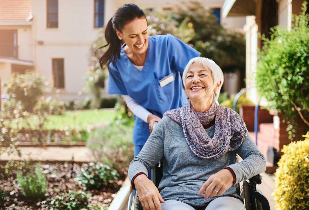 A smiling senior woman being pushed on a garden path in a wheelchair by a smiling healthcare worker.